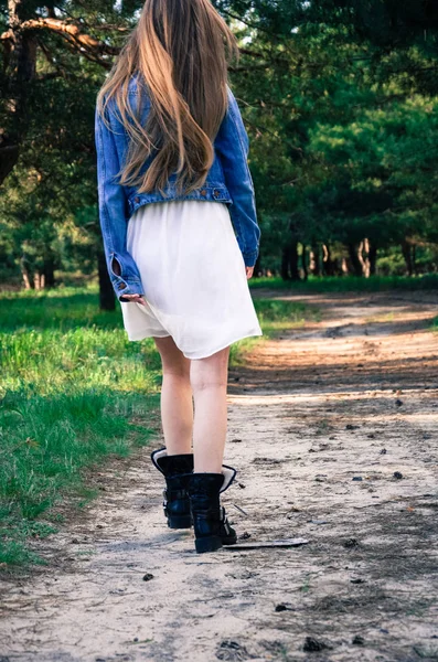 那个女孩在乡村路上走在树林里 身穿白色连衣裙和蓝色牛仔裤夹克 身穿黑色靴子的金发女郎 从后面查看 — 图库照片