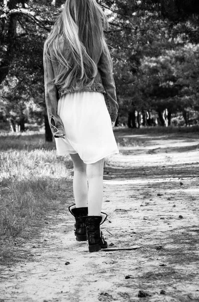 The girl walking on the contryside road in wood. Young woman with long blond hair dressed in white dress and blue jeans jacket and wearing black boots. View from back. Outdoor. Walk. Black and white.