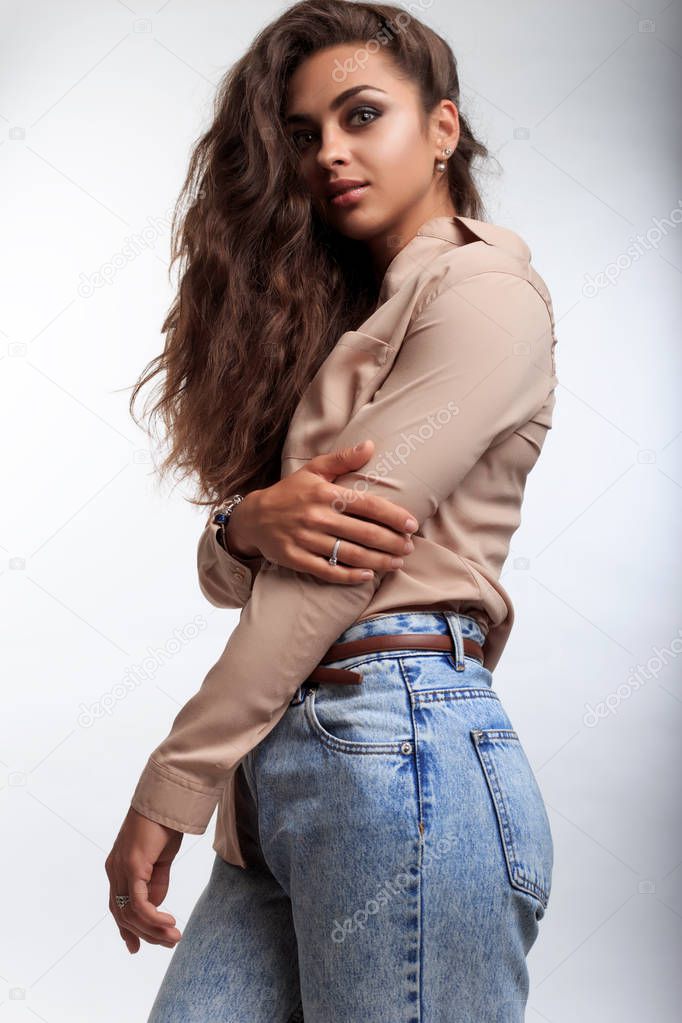 Young woman with brown skin and with very pretty face posing on the camera in studio on the white background Model standing sideways and looking at camera. Woman dressed in jeans and in shirt.