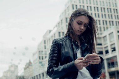 Şehirde akıllı telefon kullanan bir kadın. Deri ceketli, uzun saçlı güzel bir kadın. 
