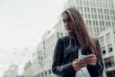 Şehirde akıllı telefon kullanan bir kadın. Deri ceketli, uzun saçlı güzel bir kadın. 