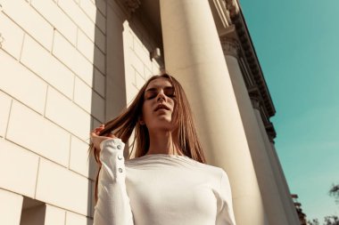 yüksek sütunlar, gökyüzü ve bir tuğla duvar arka plan karşı sarı uzun saçlı güzel bir genç kadının yatay moda portresi. Kız kameraya bakıyor. Düşük açı atışı. Güneş ışığı