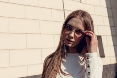 Beyaz tuğla duvara poz veren kız. Güzel genç kadın beyaz bir svidshot giyiyor ve güneş gözlüğü takıyor