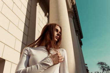 yüksek sütunlar, gökyüzü ve bir tuğla duvar arka plan karşı sarı uzun saçlı güzel bir genç kadının yatay moda portresi. Kız kameraya bakıyor. Düşük açı atışı. Güneş ışığı