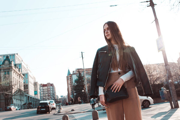 молодая девушка на фоне заходящего солнца, гуляющая по городу. Модная и стильная женщина с светлыми волосами одета в кожаную куртку и держит в руках кожаную сумку
.