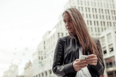Kentin arka planında siyah deri ceket ve beyaz gömlek giyen kendine güvenen bir kadın cep telefonu tutuyor..