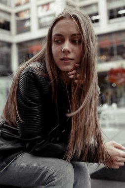 Genç kadın düşünüyor. Siyah deri ceketli kadın model. Bulanık bir binanın arkasındaki bankta oturan düz sarı saçlı kız.