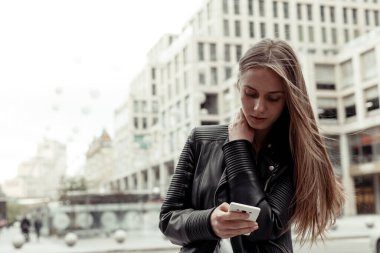 Sarışın bir kadın arka planda cep telefonuna mesaj yazıyor. Siyah deri ceket ve beyaz gömlek giyen genç bir kadın portresi. Açık hava..