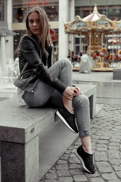 Dışarıdaki kameraya poz veren kadın model. Binanın arka planında bir bankta oturan genç sarışın kadın.