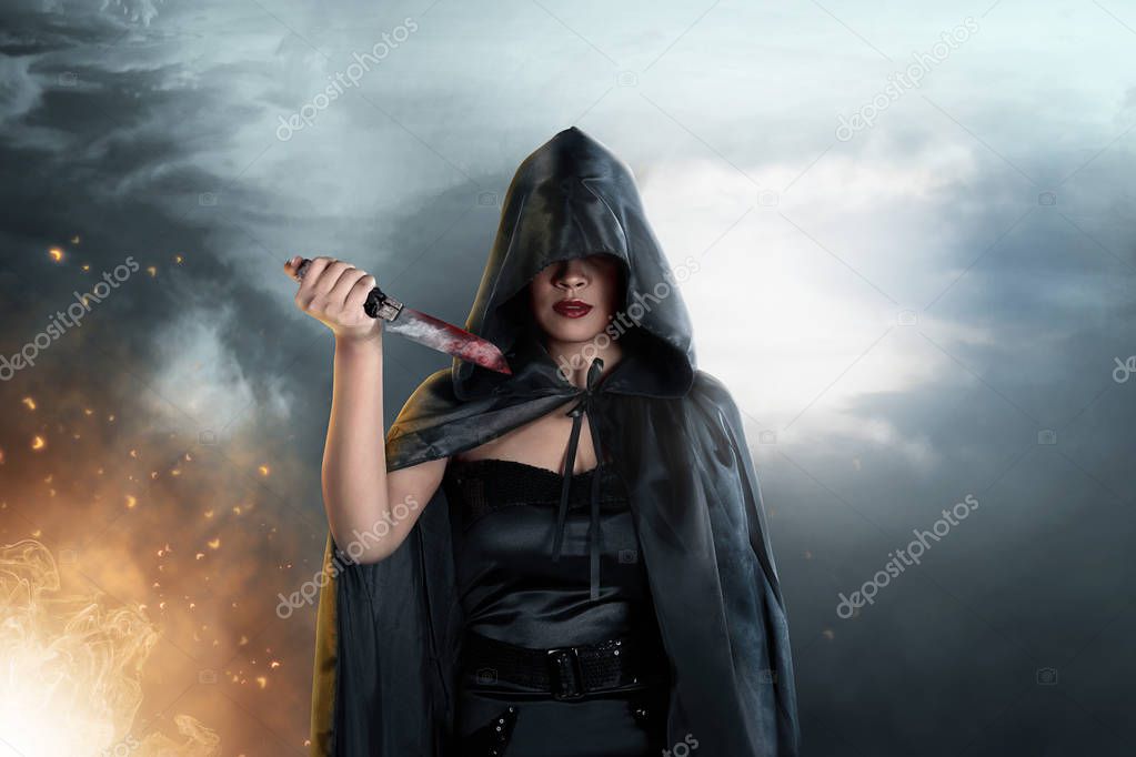 Dark witch with black hooded cloak and knife in her hands. Halloween concept
