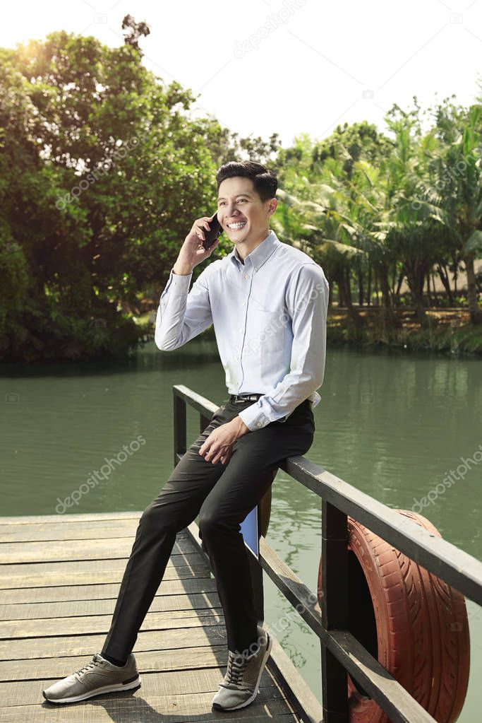 Smiling asian businessman standing while talking on the phone on the park
