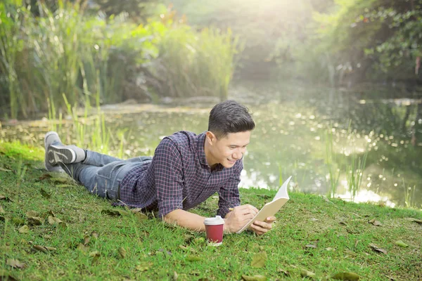 Happy asian man lying on the grass and write something on the book at park