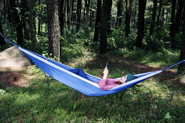 美丽的亚洲旅行妇女躺在与智能手机在吊床上的森林 — 图库照片