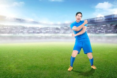 Yakışıklı Asya futbol oyuncusu mavi üniforma Stadyumu arka plan üzerinde zafer kutluyor