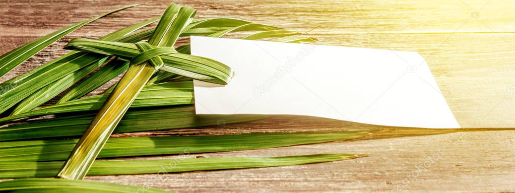 Closeup view of cross shape of palm leaf and palm branches with white blank paper and ray in wooden background. Palm Sunday concept