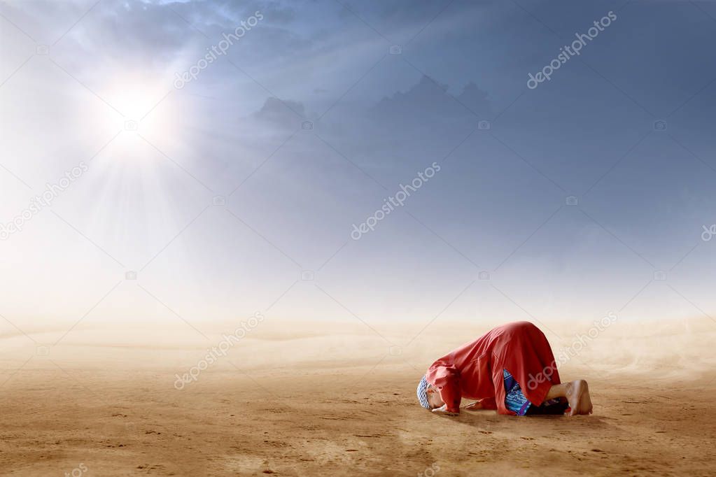 Rear view of asian muslim man praying in prostration position on desert with sun rays and dark sky background