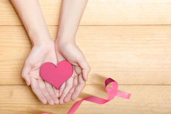 Mani umane che mostrano un cuore rosa con un nastro rosa su un legno — Foto Stock