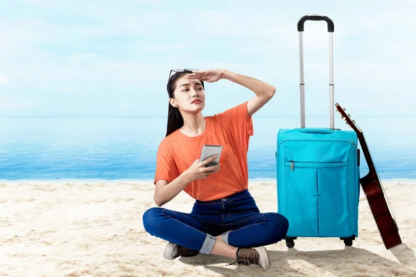 亚洲女人拿着手提箱坐在那里 手里拿着手机 望着沙滩上蓝天背景的风景 — 图库照片