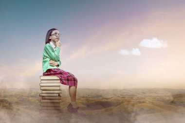 Gözlüklü Asyalı küçük kız kitap yığınının üzerinde oturuyor. Okul konseptine dönüş