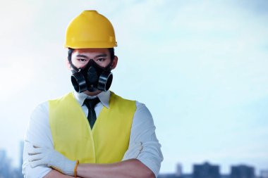 Şapkalı ve güvenlik maskeli Asyalı inşaat işçisi çapraz kollarla ayakta duruyor. İşçi Bayramı kutlu olsun.