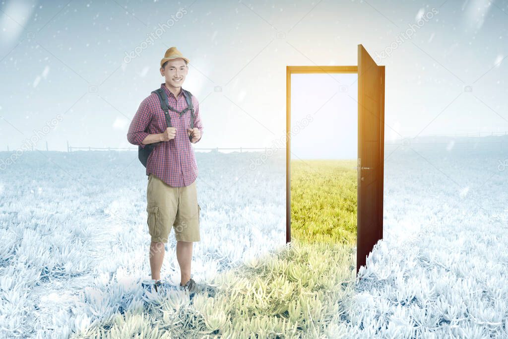 Asian man with backpack standing in front of the opened door from winter to summer climate on the meadow field. Concept of changing the environment 