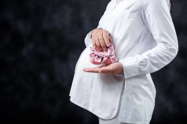 Schwangere Hält Babyschuhe Auf Dem Bauch Mit Schwarzem Hintergrund — Stockfoto