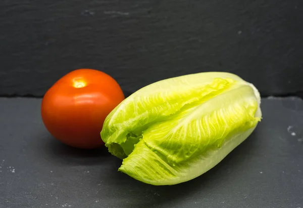 One green lettuce and two red tomatoes on a dark slate background. Focus is sharp at front of lettuce and falls off through the image.