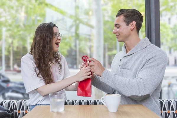 This is for you. Happy man is giving present for his loving wife. Woman is looking with excitement and smiling. Lovers are sitting at table in a restaurant