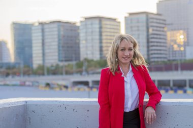 Sarışın işkadını kırmızı ceket ve siyah gömlek ayakta şehirde kameraya bakarken giyiyor