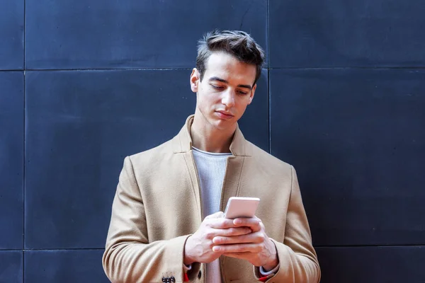 Young man wearing winter clothes in the street using the phone. Young guy with modern hairstyle with coat, blue jeans and t-shirt.