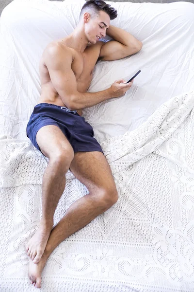 Naked dark-hair handsome man shirtless in white bed using phone