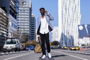 Zarif ceketli ve güneşli bir günde telefon kullanırken bir ihale sokakta yürüyen siyah Afrika genç adam önden görünümü