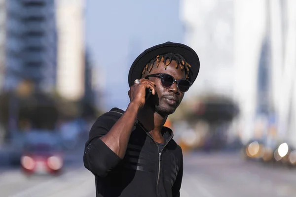 Вид спереду чорний чоловік з сонцезахисними окулярами та капелюхом, що стоїть навпроти міського пейзажу на вулиці, використовуючи мобільний телефон у сонячний день . — стокове фото