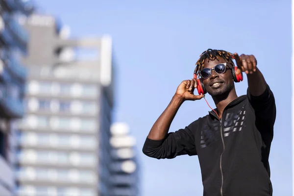 Rahat kıyafetler ve güneşli bir günde müzik dinlemek için kulaklık kullanırken sokağın ortasında duran güneş gözlüğü takan bir zenci önden görünümü