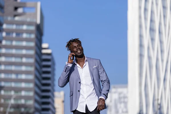 Вид спереду усміхненого молодого чорношкірого африканця в елегантному одязі, що стоїть на вулиці, використовуючи мобільний телефон на відкритому повітрі в сонячний день — стокове фото