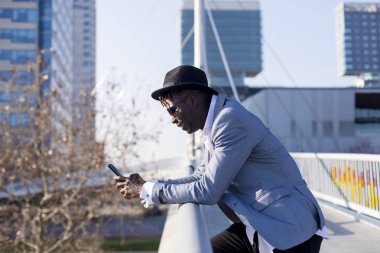 Siyah Afrika genç adam açık havada güneşli bir günde bir cep telefonu kullanırken bir metal çit yaslanmış güneş gözlüğü ve şapka giyen yan görünüm