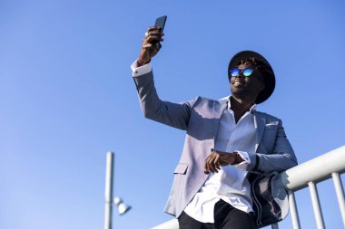şapka ve güneş gözlüğü üzerinde mavi gökyüzü güneşli bir günde bir selfie çekerken rahatlatıcı bir metal çit yaslanmış giyen genç Afrika siyah adam düşük açılı görünümü