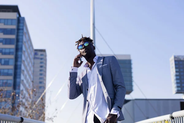 Вид спереду усміхненого молодого чорношкірого африканця в елегантному одязі, що стоїть на вулиці, використовуючи мобільний телефон на відкритому повітрі в сонячний день — стокове фото