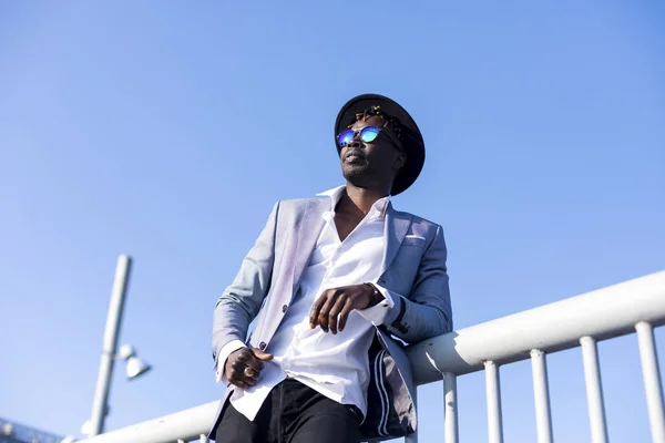 Низький кут зору молодого африканського чорного чоловіка в капелюсі та сонцезахисних окулярах, що спираються на металевий паркан, розслабляючись, дивлячись в сонячний день на блакитне небо — стокове фото