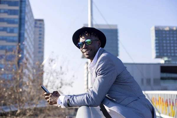 Вид збоку чорношкірого африканського молодого чоловіка в капелюсі і сонцезахисних окулярах, що спираються на металевий паркан, використовуючи мобільний телефон на відкритому повітрі в сонячний день — стокове фото