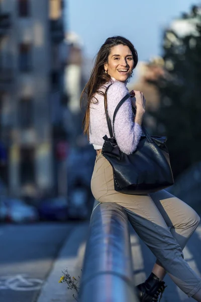 Vue de face de la belle jeune femme tendance portant des vêtements décontractés assis sur une clôture métallique dans la rue tout en regardant loin par une journée ensoleillée — Photo