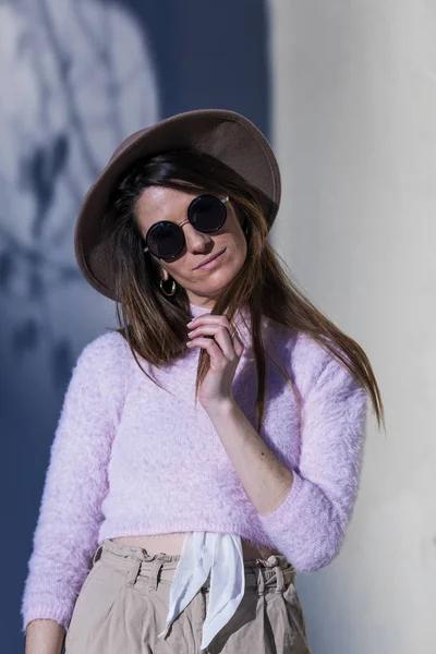 Přední pohled na krásné moderní mladé ženy, které nosí sluneční brýle a klobouk postával na ulici a pózuje pro kamery v slunečný den — Stock fotografie