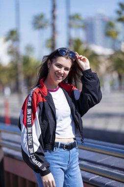 Güzel gülümseyen genç kadın kentsel giyim ve kamera açık havada sokakta parlak bir günde bakarken ayakta güneş gözlüğü takmış önden görünümü