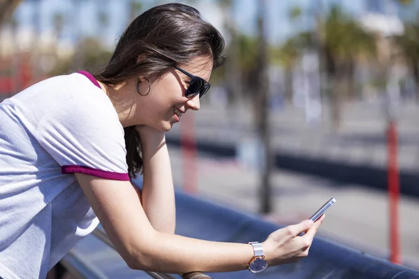 Vista lateral de una hermosa mujer joven con ropa urbana apoyada en una valla metálica mientras usa un teléfono móvil al aire libre en la calle en un día brillante — Foto de Stock
