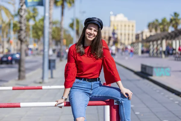 Vista frontal de una hermosa y sonriente joven vestida con ropa urbana sentada en una valla metálica mientras mira la cámara al aire libre en la calle en un día brillante — Foto de Stock