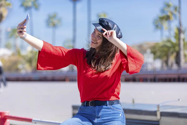 Vista frontal de una joven sonriente ropa casual sentada en una valla metálica mientras toma una selfie en un día brillante — Foto de Stock