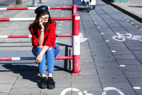 Vista frontal de una hermosa joven vestida con ropa urbana sentada en una valla metálica mientras usa un teléfono móvil al aire libre en la calle en un día brillante — Foto de Stock