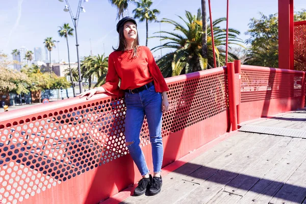 Vista frontal de una hermosa joven vestida con ropa urbana apoyada en una valla de puente metálico mientras mira al aire libre en un día brillante — Foto de Stock