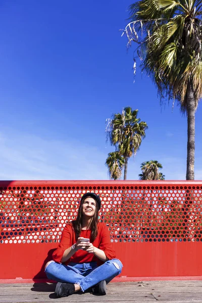 Vista frontal de una hermosa joven vestida con ropa urbana sentada en el piso de un puente mientras usa un teléfono móvil al aire libre en un día brillante — Foto de Stock