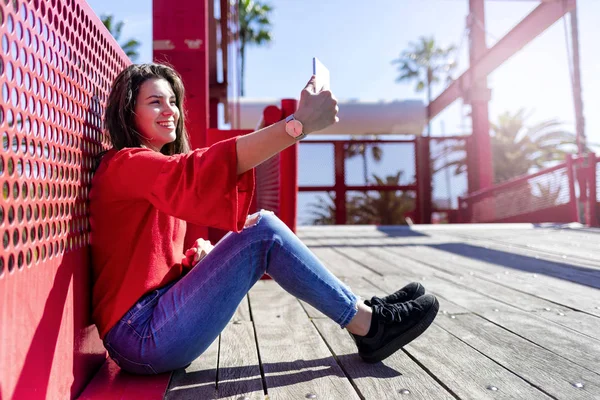 Vista frontal de una hermosa y sonriente joven vestida con ropa urbana sentada en el suelo mientras toma una selfie y hace un gesto al aire libre en la calle en un día brillante — Foto de Stock